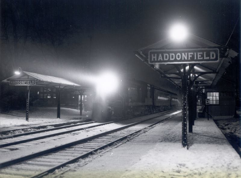 Black-and-white night photo of the Haddonfield train platform in falling snow under a lamp