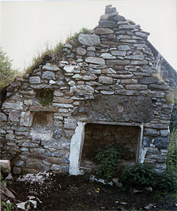A ruined stone cottage with an open hearth and a tall gable, grass growing over the walls