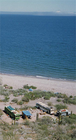 Buses and tents on a sandy shore above the clear blue water of a bay, seen from a hill