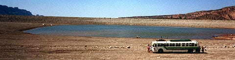 A bus, small in the distance, on a dry lakeshore below red bluffs
