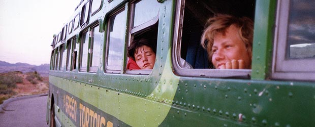 Two passengers smiling out of the windows of the green bus on a desert road