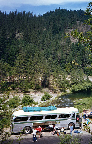 A Green Tortoise bus parked beside a creek below forested hills, passengers on the bank