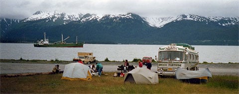 Tents and a bus on a gravel shore, a ship and snow-capped mountains across the water